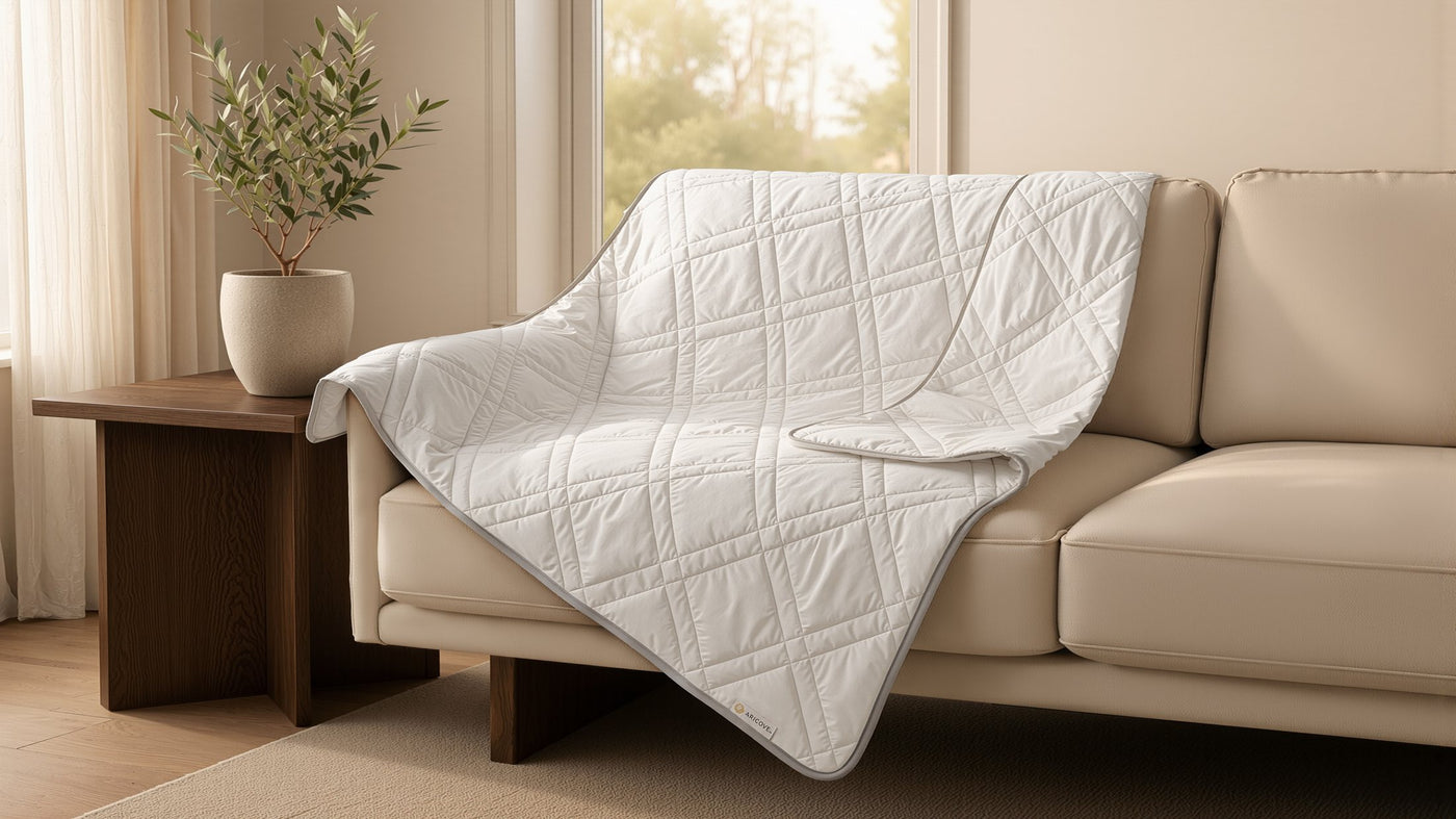 White quilted Aricove weighted blanket draped over a cream sofa, with a wooden side table and small olive tree to the left, soft daylight from a window behind, Aricove label visible at the blanket corner